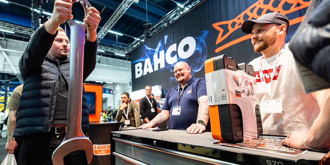 Why exhibit - Exhibitors and visitors meet at a trade show booth to discuss products, with a focus on networking and collaboration. Visitor holding a giant wrench. How long can he hold it?  